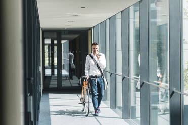 Smiling businessman talking on smart phone and walking with bicycle on footbridge - UUF30921