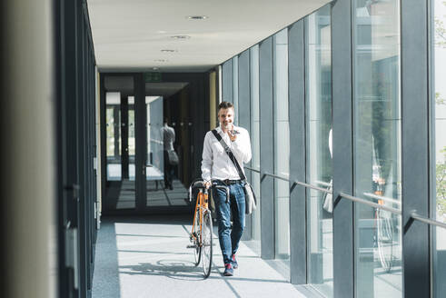 Lächelnder Geschäftsmann, der mit seinem Smartphone telefoniert und mit dem Fahrrad auf einer Fußgängerbrücke spazieren geht - UUF30921