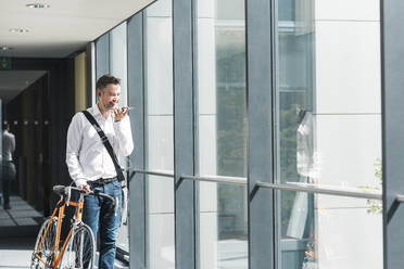 Lächelnder Geschäftsmann, der mit einem Smartphone spricht und mit einem Fahrrad am Fenster steht - UUF30922