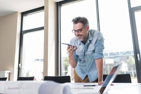 Zufriedener Architekt, der im Büro mit seinem Smartphone über einen Bauplan diskutiert - UUF30928