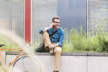 Happy businessman sitting and talking on smart phone at office park - UUF30934