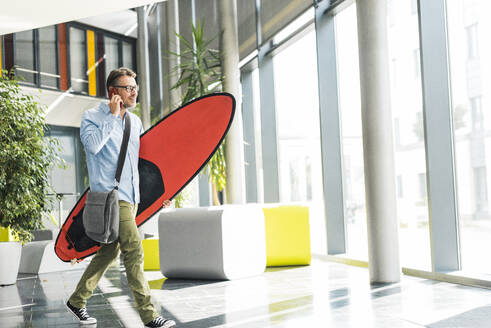 Geschäftsmann spricht mit Smartphone und trägt Surfbrett im Büro - UUF30936