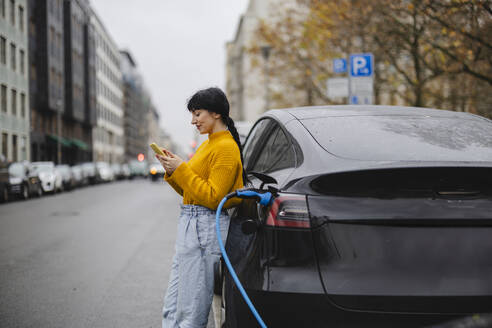 Frau benutzt Smartphone in der Nähe eines Elektroautos auf der Straße - JCCMF11066