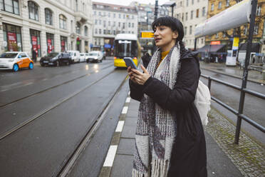 Lächelnde Frau mit Smartphone an der Straßenbahnhaltestelle - JCCMF11091