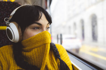 Frau, die ihr Gesicht mit einem gelben Pullover bedeckt und über drahtlose Kopfhörer im Bus Musik hört - JCCMF11100