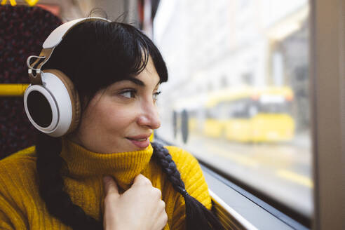 Smiling woman wearing wireless headphones sitting near window in bus - JCCMF11101