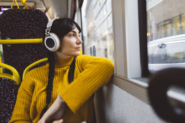 Thoughtful woman wearing wireless headphones and sitting in bus - JCCMF11103