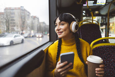 Glückliche Frau mit drahtlosen Kopfhörern und Kaffeetasse mit Smartphone im Bus - JCCMF11106