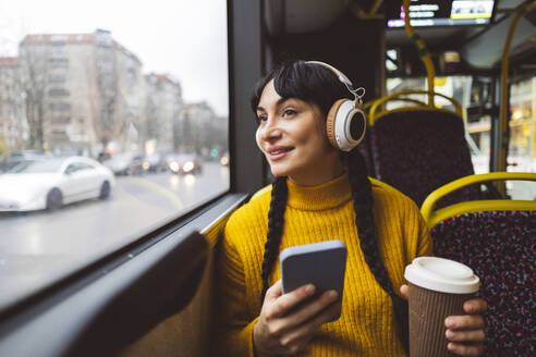 Happy woman wearing wireless headphones and holding coffee cup with smart phone in bus - JCCMF11106
