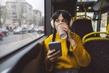 Frau trägt drahtlose Kopfhörer und trinkt Kaffee im Bus - JCCMF11107