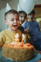 Junge mit offenem Mund beim Ausblasen von Geburtstagskerzen auf Kuchen zu Hause - ANAF02633
