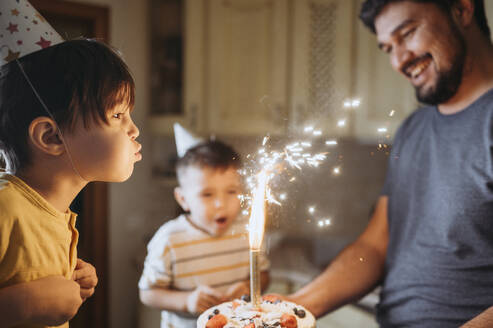 Boy wearing party hat and blowing candle on cake with family - ANAF02639