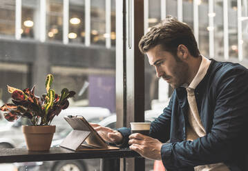 Young businessman holding disposable cup and using tablet PC at cafe - UUF30940