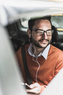 Smiling businessman wearing in-ear headphones and sitting in car - UUF30953