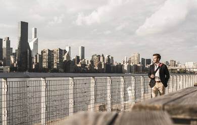 Geschäftsmann, der in der Nähe des Geländers am Hudson River in New York City, USA, spazieren geht - UUF30956