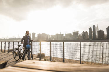 Businessman pushing bicycle by river at sunset in New York City - UUF30966