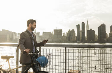 Lächelnder Geschäftsmann, der ein Fahrrad schiebt und sein Smartphone am Fluss in New York City benutzt - UUF30967