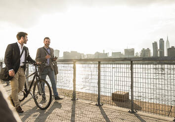 Geschäftsmann, der ein Fahrrad schiebt und mit einem Kollegen am Fluss in New York City, USA, spazieren geht - UUF30969