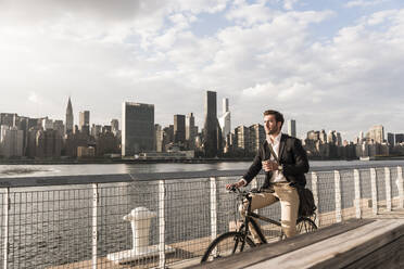 Businessman holding coffee cup and cycling by river in New York City, USA - UUF30971