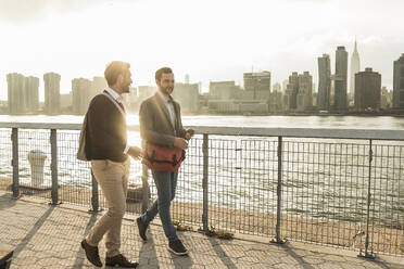 Happy young colleagues talking by river at sunset in New York City, USA - UUF30974