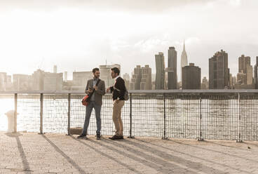 Young colleagues leaning on railings and talking by river at sunset in New York City - UUF30975