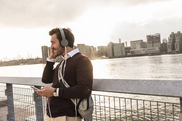 Smiling businessman wearing headphones and listening to music by Hudson river at sunset in New York City - UUF30979