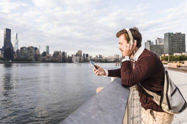 Young businessman wearing headphones and listening to music by Hudson river in New York City, USA - UUF30980