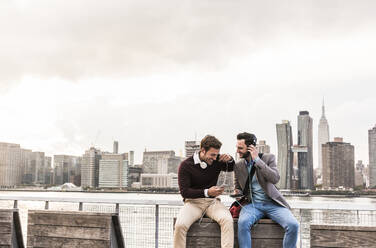 Happy business colleagues wearing headphones and listening to music in front of New York City skyline - UUF30982