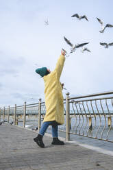 Woman standing near railing and reaching at seagulls flying near sea - OLRF00088