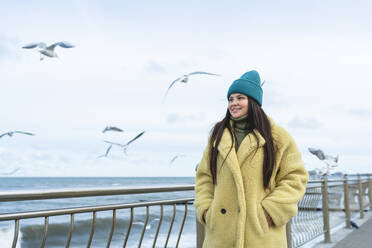 Lächelnde Frau mit Händen in den Taschen, die in der Nähe des Geländers an der Promenade spazieren geht - OLRF00091