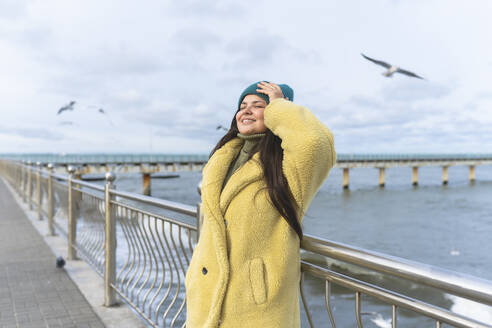 Smiling woman with head in hands leaning on railing near sea - OLRF00093