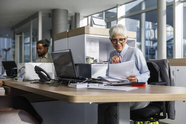 Lächelnde ältere Geschäftsfrau, die am Schreibtisch sitzend ein Dokument liest - IKF01520
