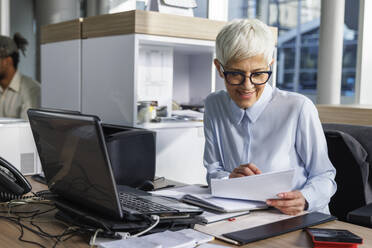 Smiling businesswoman examining document sitting at desk in office - IKF01521