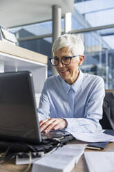 Lächelnde ältere Geschäftsfrau bei der Arbeit am Laptop am Schreibtisch im Büro - IKF01525