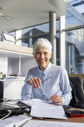 Happy senior businesswoman holding eyeglasses sitting at desk in office - IKF01527