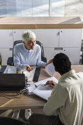 Geschäftsfrau zeigt einem Kollegen, der am Schreibtisch sitzt, Dokumente im Büro - IKF01533