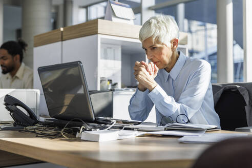 Worried businesswoman with hands clasped looking at laptop in office - IKF01559