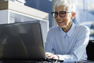 Glückliche Geschäftsfrau, die eine Brille trägt und einen Laptop im Büro benutzt - IKF01564