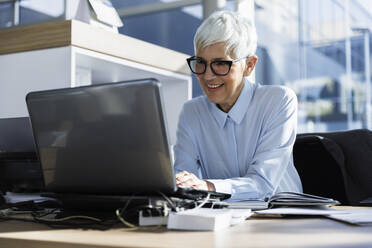 Lächelnde ältere Geschäftsfrau mit Laptop am Schreibtisch sitzend im Büro - IKF01565