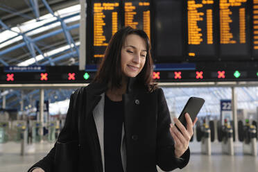 Lächelnde reife Frau, die am Bahnhof ein Mobiltelefon benutzt - ASGF04809