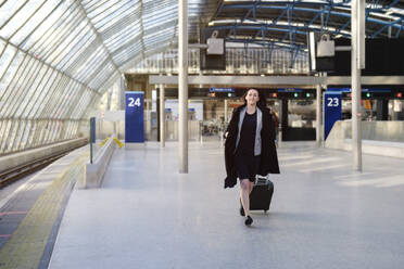 Smiling mature woman walking with suitcase on railway station platform - ASGF04812
