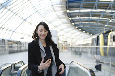 Glückliche reife Frau mit Smartphone am Bahnhof - ASGF04817