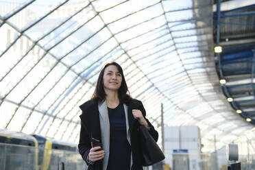 Lächelnde reife Frau mit Handtasche und Smartphone am Bahnhof - ASGF04820