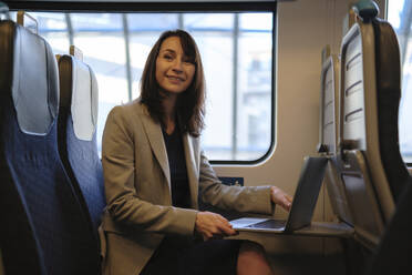 Smiling businesswoman sitting with laptop in train - ASGF04821