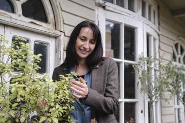 Smiling woman near plants in front of house - ASGF04832