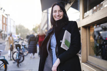 Happy woman standing with newspaper on footpath near building - ASGF04852