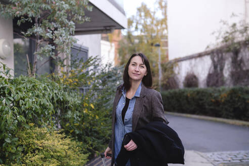 Smiling mature woman walking near plants at footpath - ASGF04856