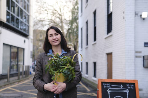 Smiling mature woman standing with potted plant near buildings - ASGF04867