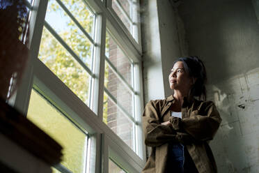 Thoughtful woman with arms crossed looking out through window at home - JOSEF22742