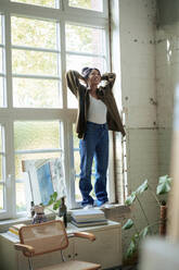 Smiling woman with hands behind head standing on window sill at home - JOSEF22744